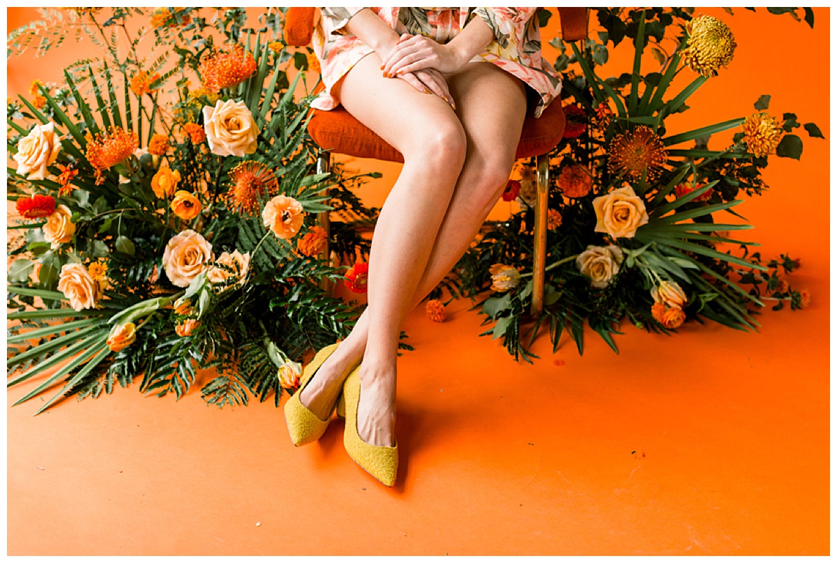 A photo-op featuring a burnt orange velvet midcentury chair set in front of a burnt orange backdrop and surrounded by a lush tropical-inspired floral inspiration. 