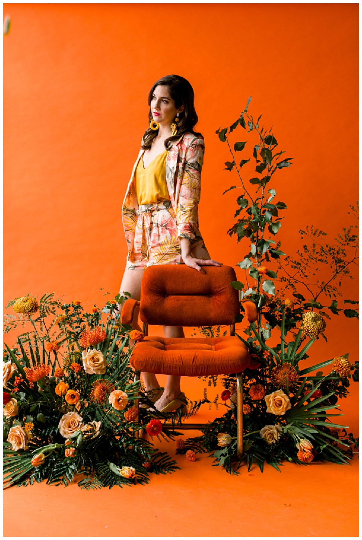 A photo-op featuring a burnt orange velvet midcentury chair set in front of a burnt orange backdrop and surrounded by a lush tropical-inspired floral inspiration. 