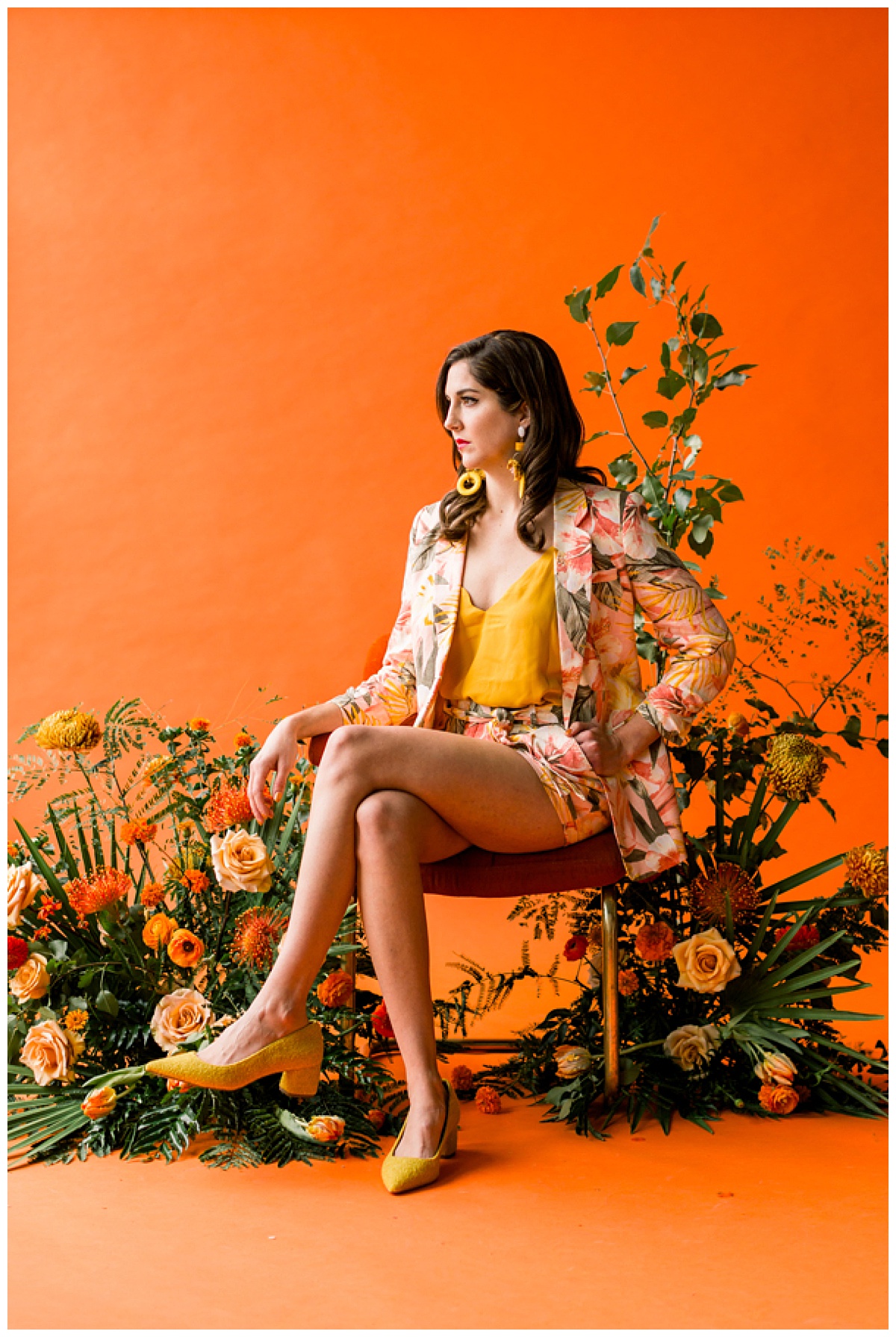 Model poses on a burnt orange velvet midcentury chair set in front of a burnt orange backdrop and surrounded by a lush tropical-inspired floral inspiration. 