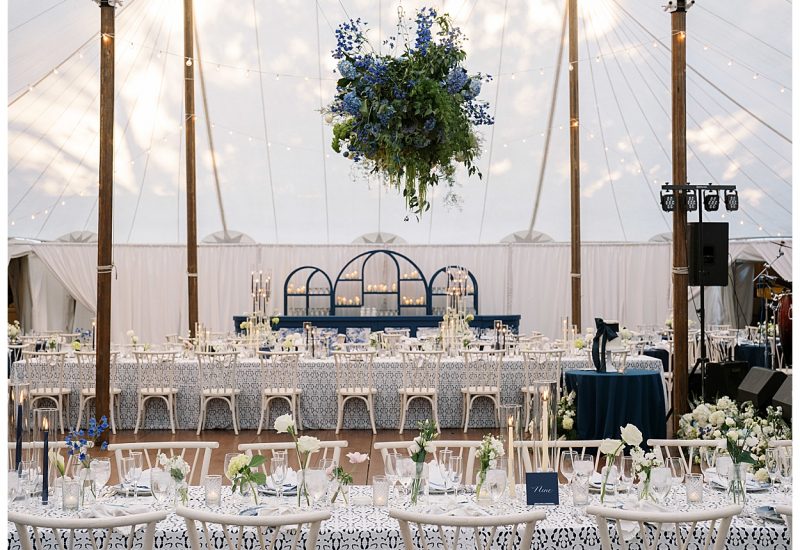 An elegant wedding reception under a tent featuring blue and white tablescapes, white chairs, and a grand bar with arches and candles creating a sophisticated atmosphere. Rentals by Something Vintage Rentals, the U.S. East Coast’s best event rental and wedding decor company.