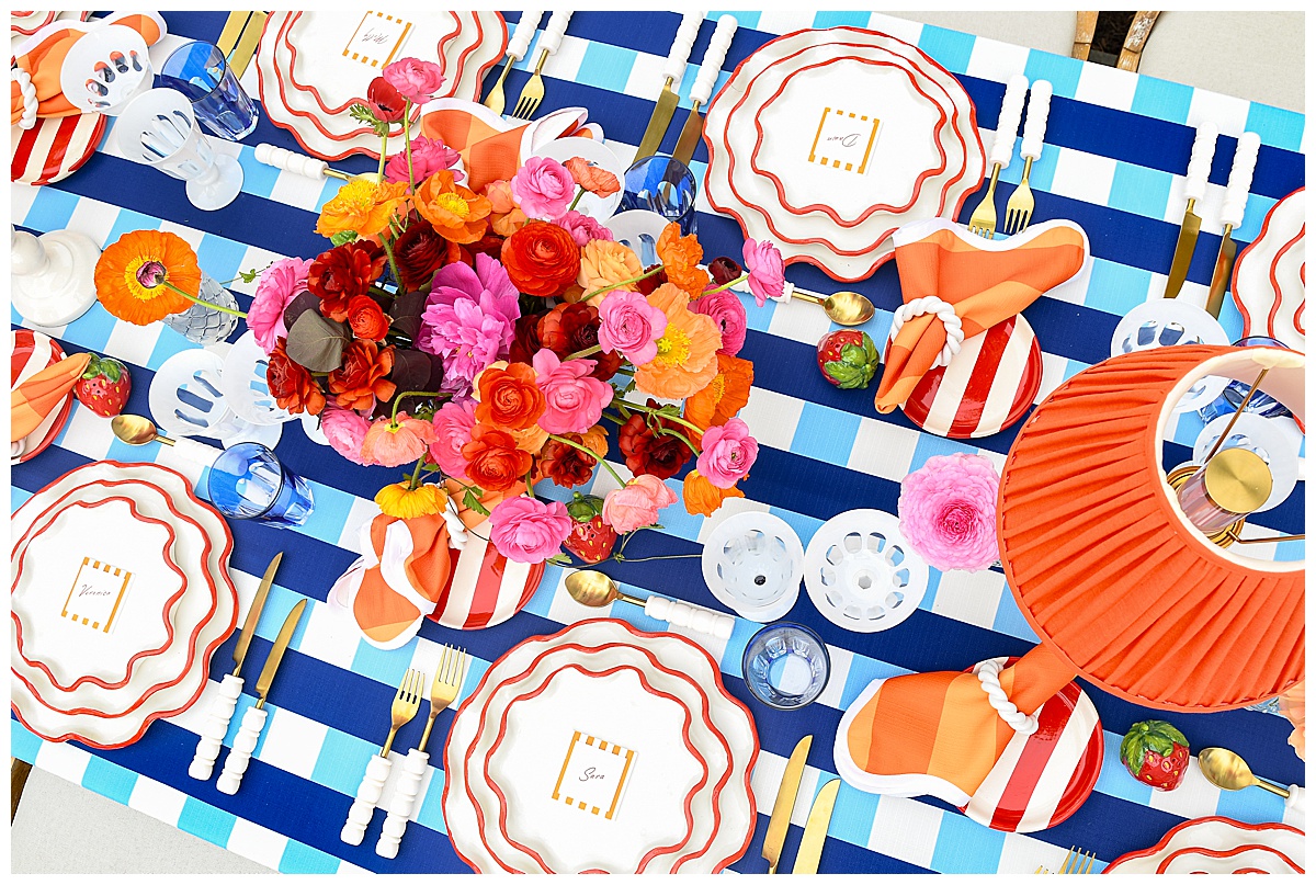 A vibrant summer tablescape with a blue and white plaid linen, red-rimmed ruffle plates, bold orange striped napkins, and a lush centerpiece of bright florals, creating a playful and colorful setting. Rentals by Something Vintage Rentals, the U.S. East Coast’s best event rental and wedding decor company.