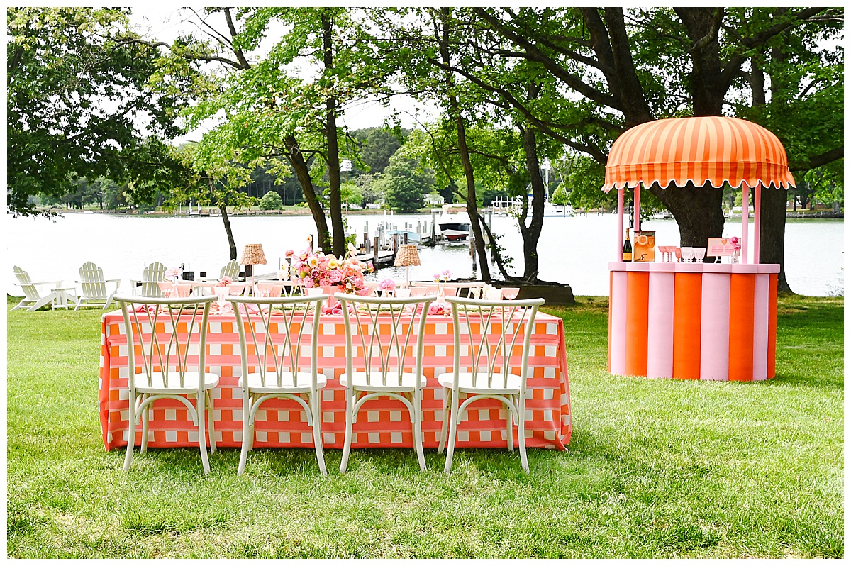 Outdoor waterfront setup with a coral checkered dining table and matching striped bar under the trees. Rentals by Something Vintage Rentals, the U.S. East Coast’s best event rental and wedding decor company.